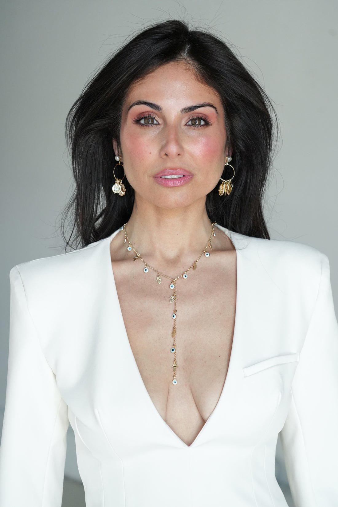A woman with dark hair wearing a white blazer and a gold lariat-style necklace with clear rhinestones, photographed against a plain light gray background.