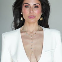 A woman with dark hair wearing a white blazer and a gold lariat-style necklace with clear rhinestones, photographed against a plain light gray background.